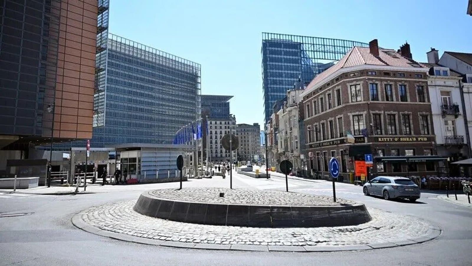 A quiet street corner in the European quarter in Brussels under a clear sky, with the Berlaymont building on the left and a parked car passing a cobbled traffic island.