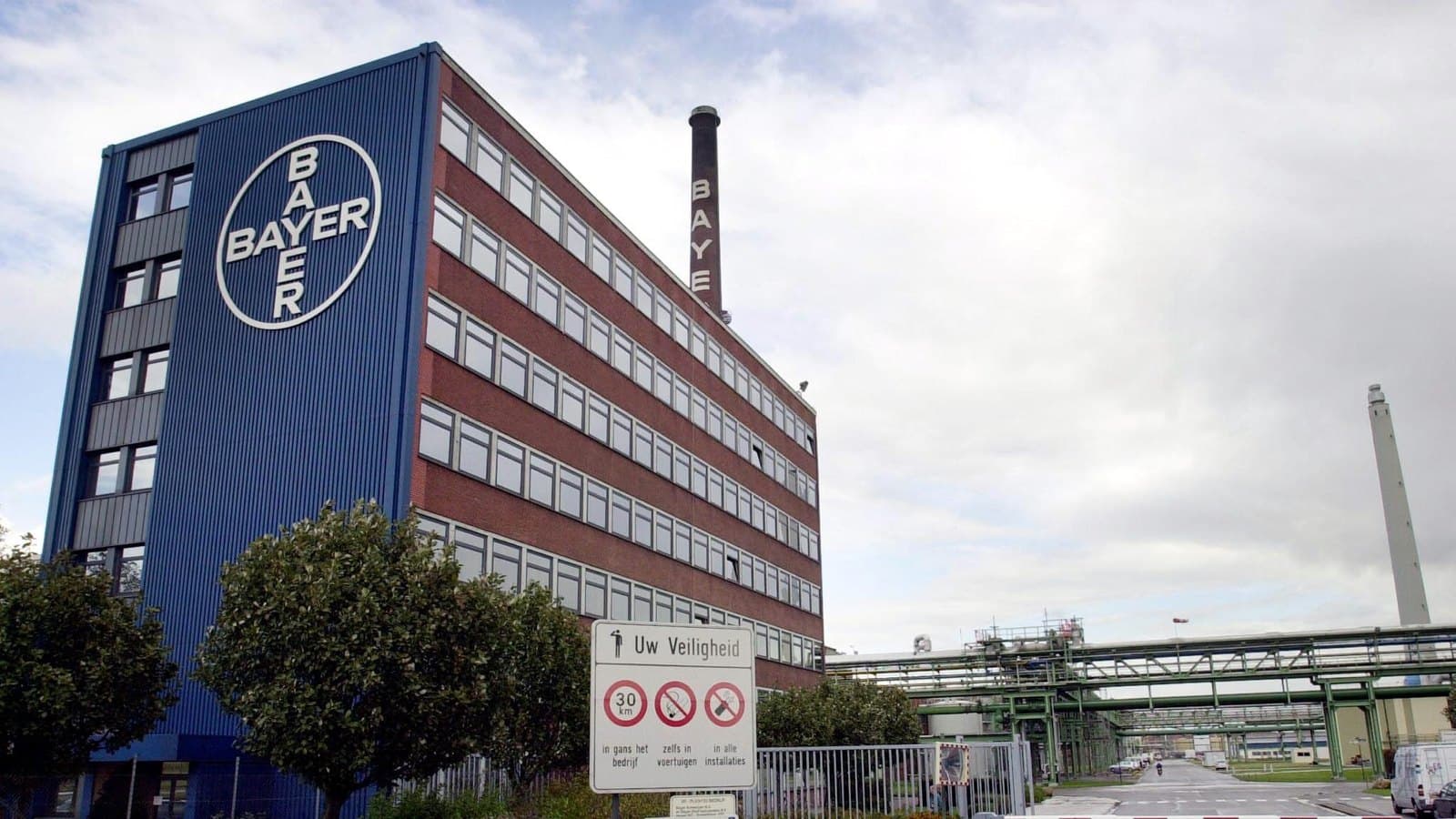 The Bayer site in Antwerp, the company name on the office block beside a chimney, with pipework and a barrier at the plant entrance.