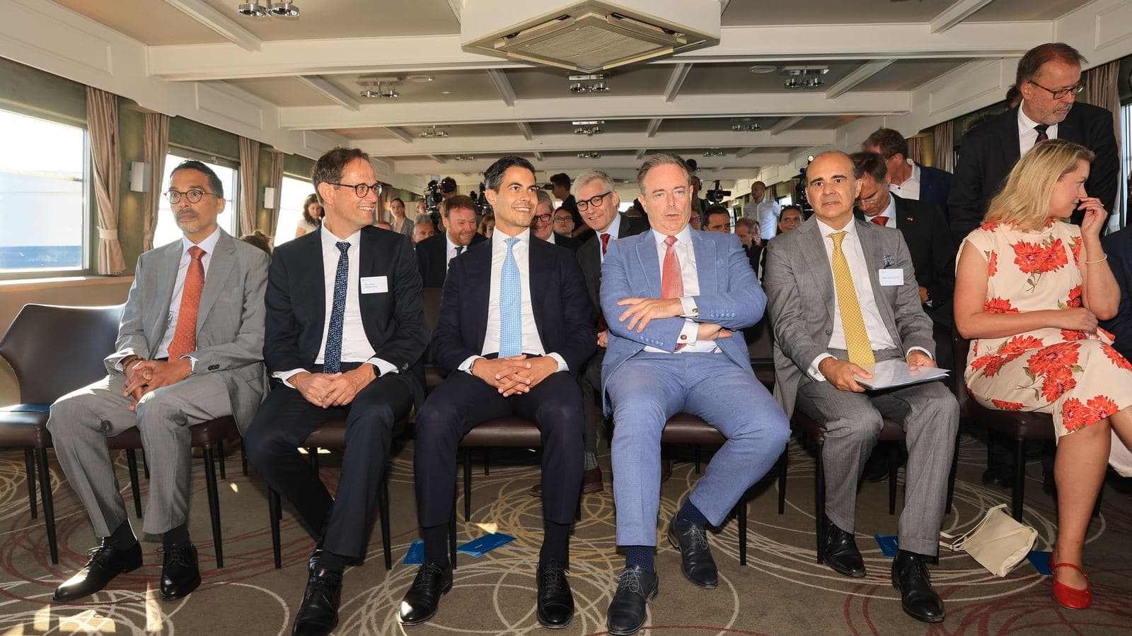 Guests and officials seated in the front row at an inauguration event in the port of Antwerp, with photographers and other attendees standing behind them.
