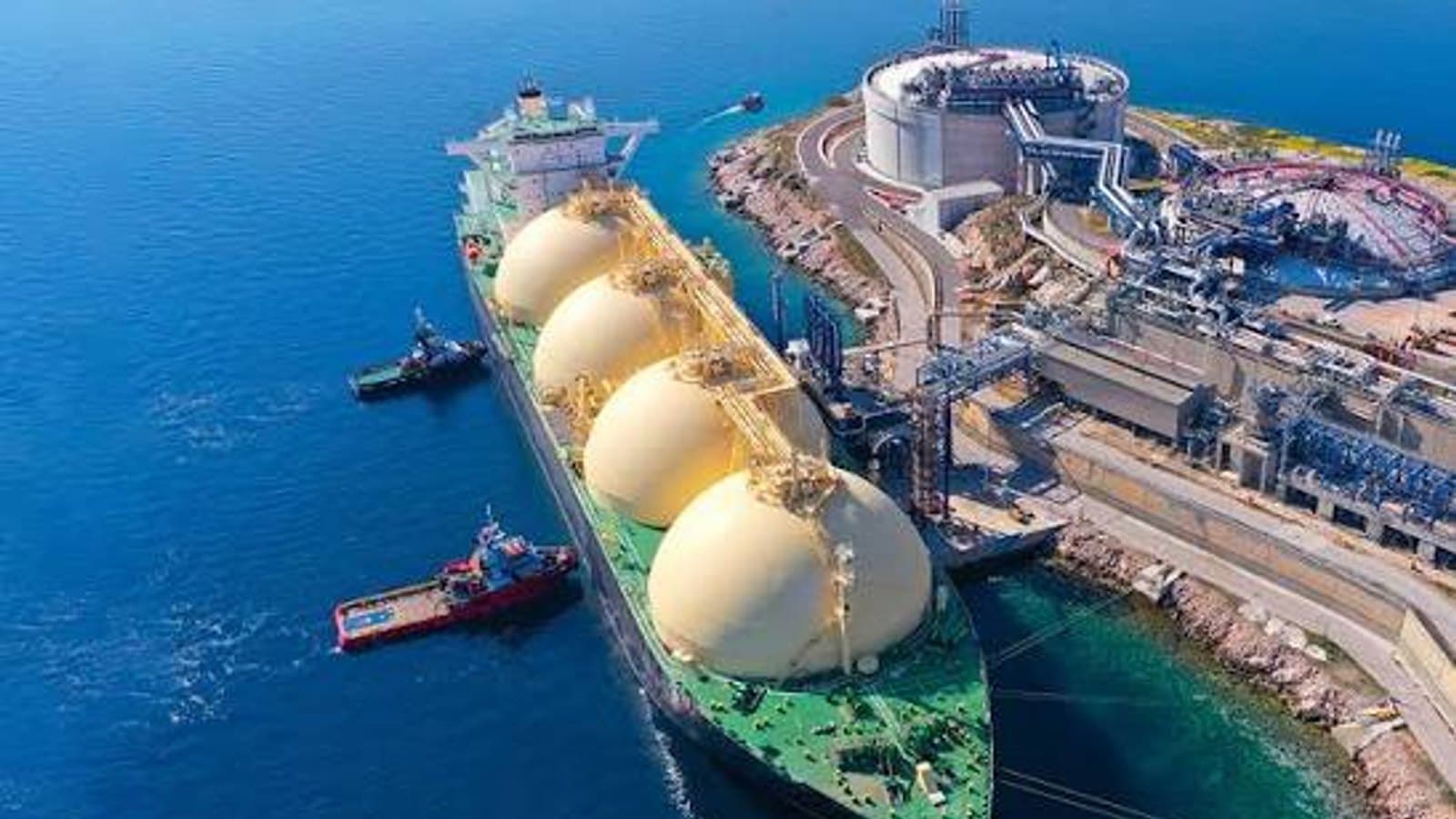 An aerial view of a liquefied natural gas carrier with spherical tanks berthed at a coastal terminal, with two tugs alongside.