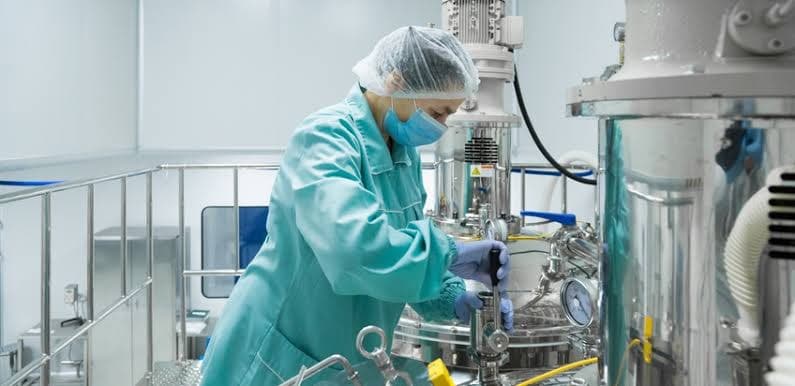 A lab technician in a hairnet and mask operates a stainless steel bioreactor at a pharmaceutical manufacturing plant