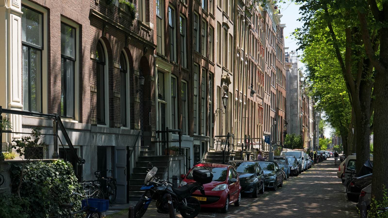 Sunlight and shadows on the old brick house facades at the street Binnenkant, with city trees along the canal border to the right; free photo Amsterdam canals, Fons Heijnsbroek Summer 2022. File photograph.