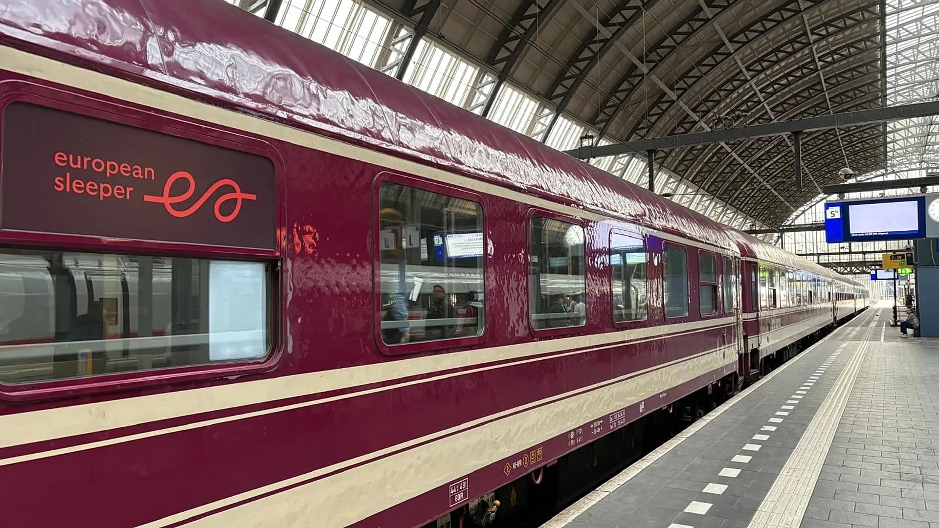 A maroon and cream European Sleeper train carriage waiting at a station platform under a glass roof