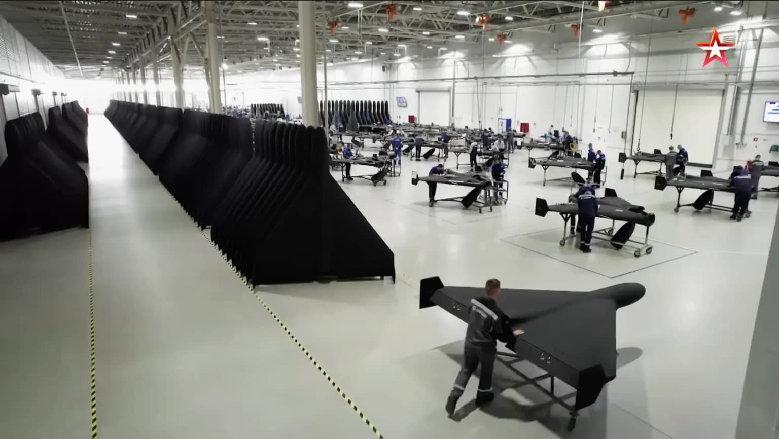 Workers assemble rows of dark attack drones on a factory floor, with a Russian state television logo visible in the corner of the screenshot