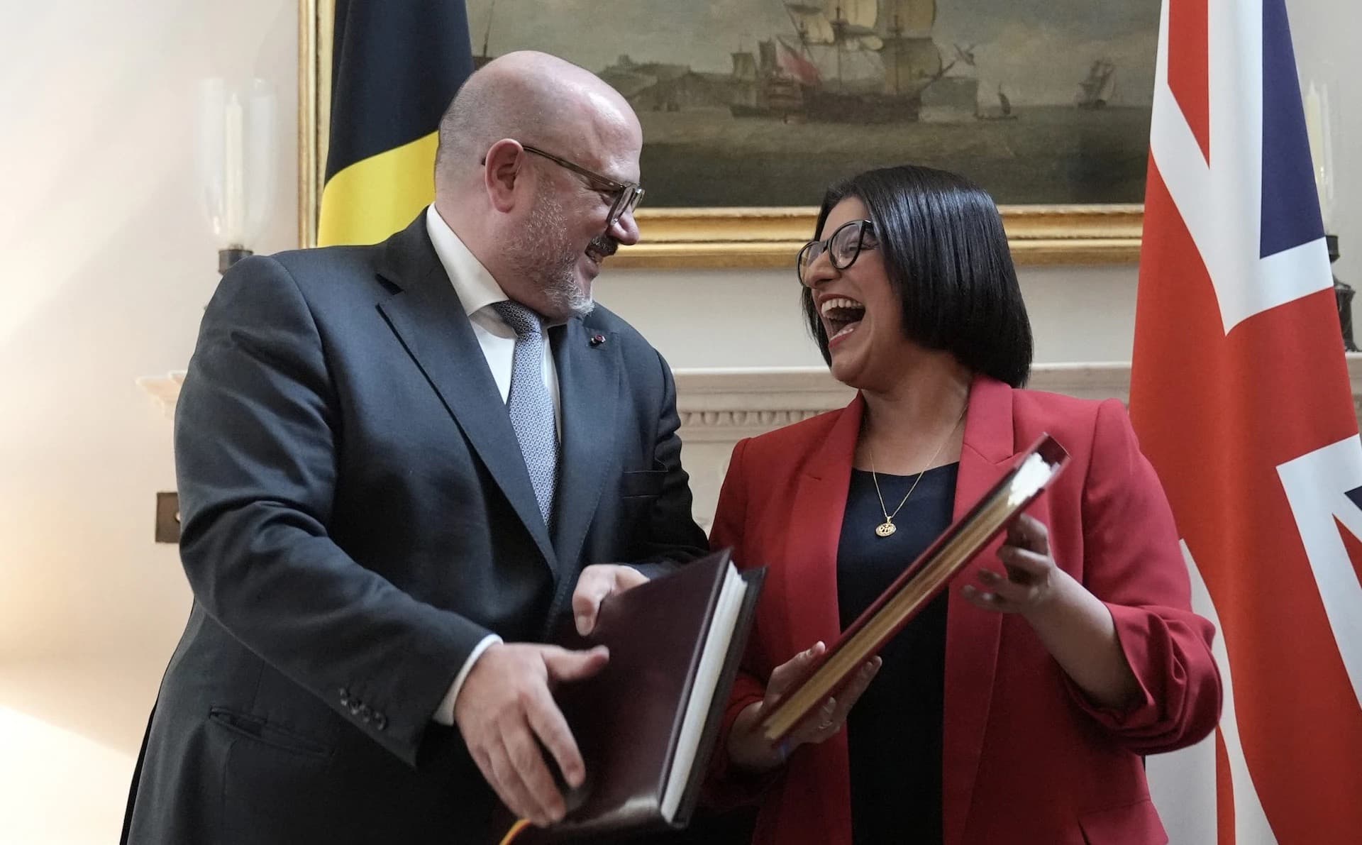 Belgian interior minister Bernard Quintin and UK Home Secretary Shabana Mahmood laughing together while exchanging signed documents in front of Belgian and British flags
