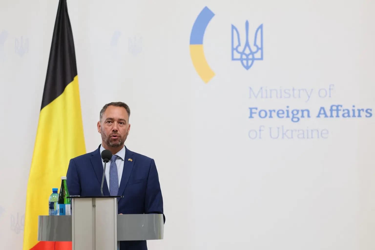 Belgian Foreign Minister Maxime Prevot speaking at a podium beside a Belgian flag and a backdrop for Ukraine's Ministry of Foreign Affairs
