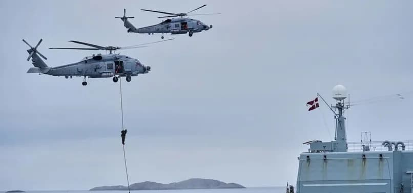 Two military helicopters hover over a naval vessel flying the Danish flag as a soldier rappels down during an Arctic exercise