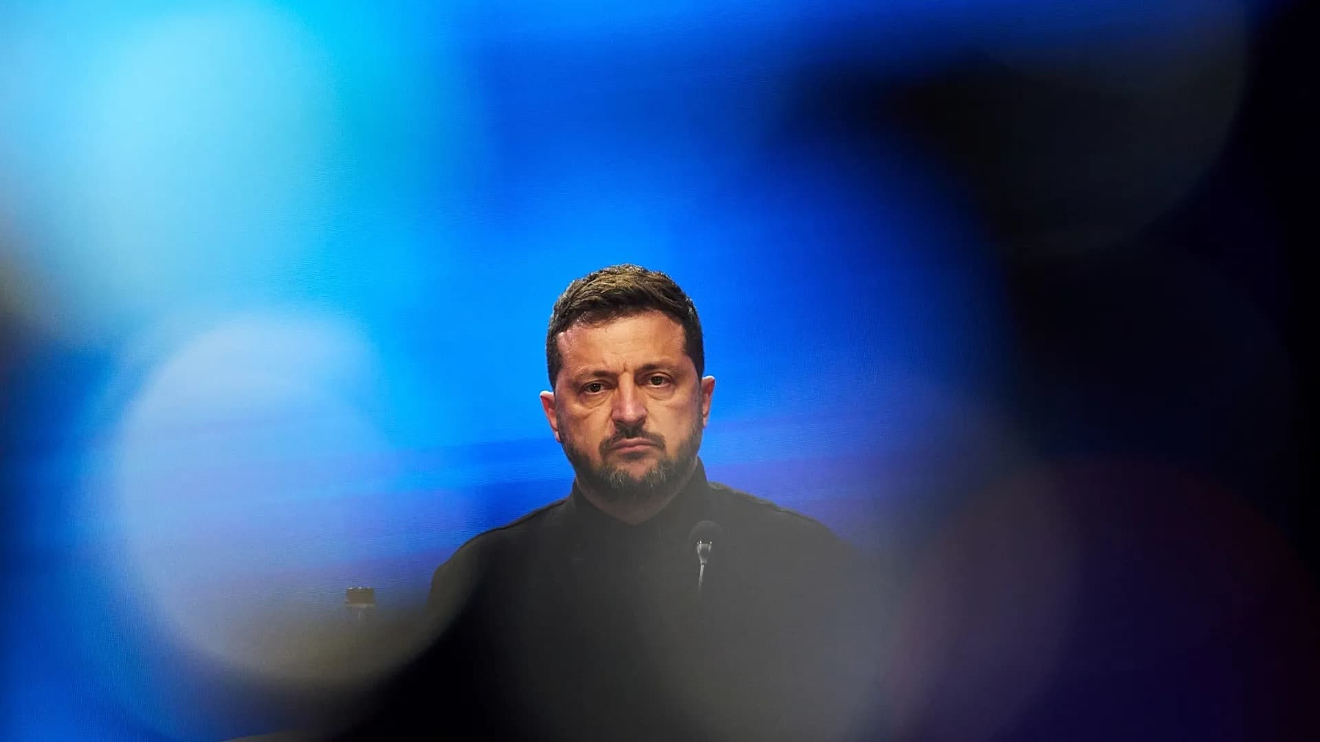 A blurred, moody portrait of Ukrainian President Volodymyr Zelensky lit in blue at a podium