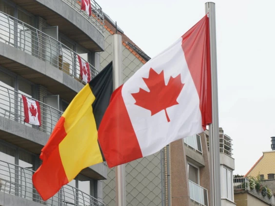 Belgian and Canadian flags fly side by side on a pole in front of an apartment building