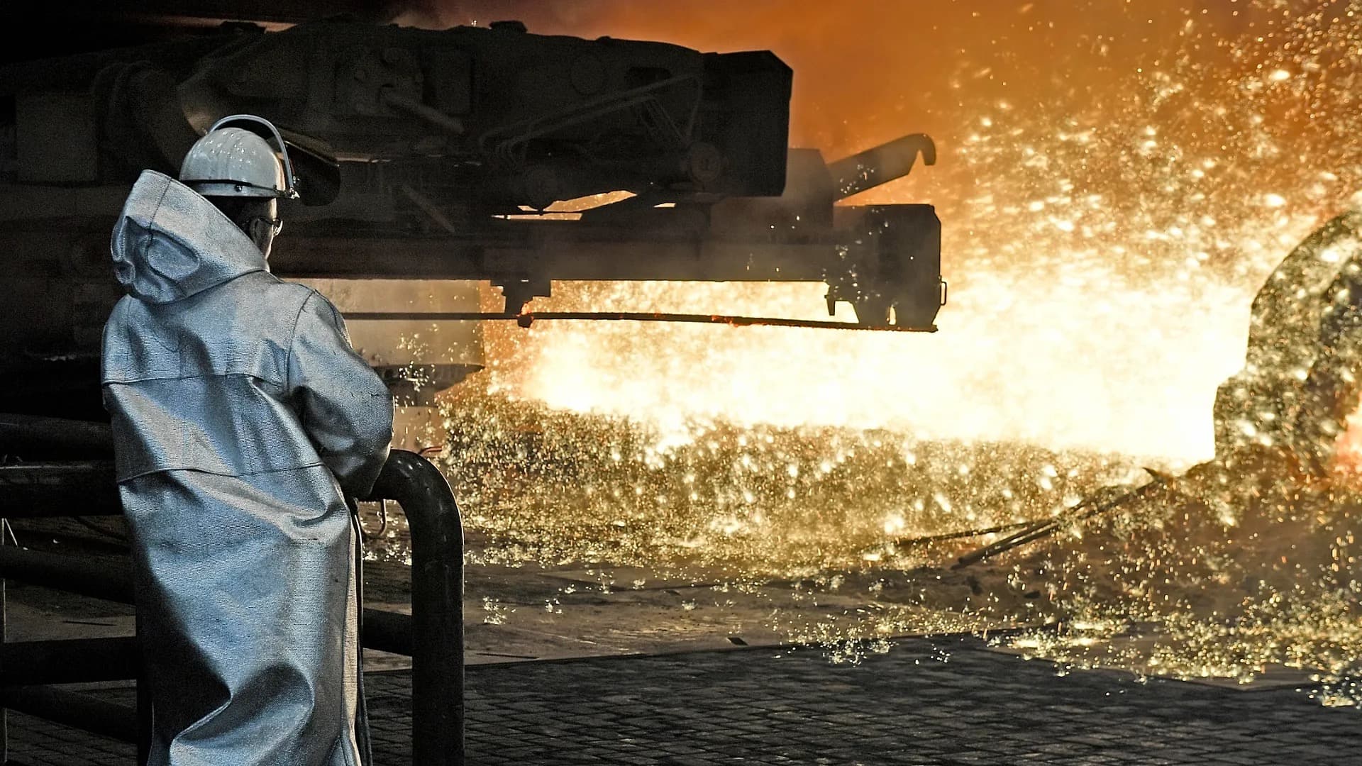 A steelworker in protective gear watching molten metal and sparks fly at a foundry