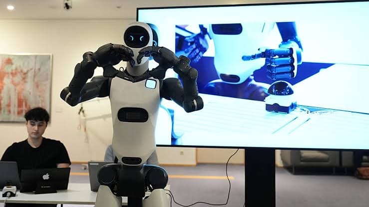 A white and black humanoid robot raising its arms during a demonstration in front of onlookers