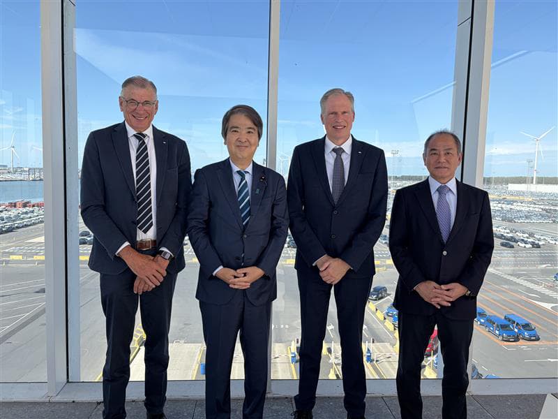 Four men in suits posing for a photo with a view of the Zeebrugge car terminal and wind turbines behind them