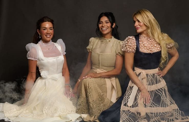 Three women in elegant dresses pose together laughing, smoke swirling around them in a studio portrait