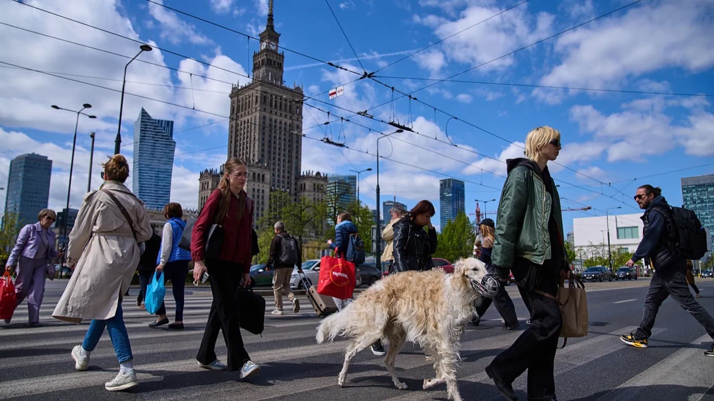 Pedestrians and a dog cross a busy street in Warsaw with the Palace of Culture and Science visible in the background